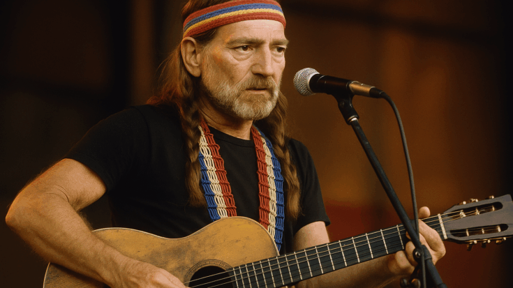Willie Nelson playing an acoustic guitar onstage, wearing a colorful headband and a beaded strap while singing into a microphone.