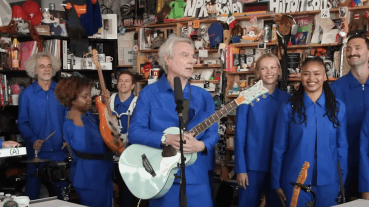 David Byrne performing his Tiny Desk Concert with his band, all dressed in blue uniforms, surrounded by NPR’s cluttered office backdrop during his “Who Is the Sky?” tour stop.