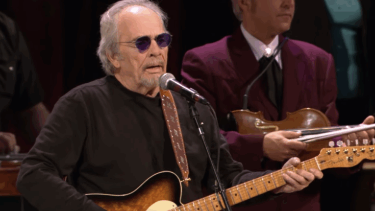 Merle Haggard performing onstage with his guitar, wearing sunglasses and backed by his band during a live country music show.